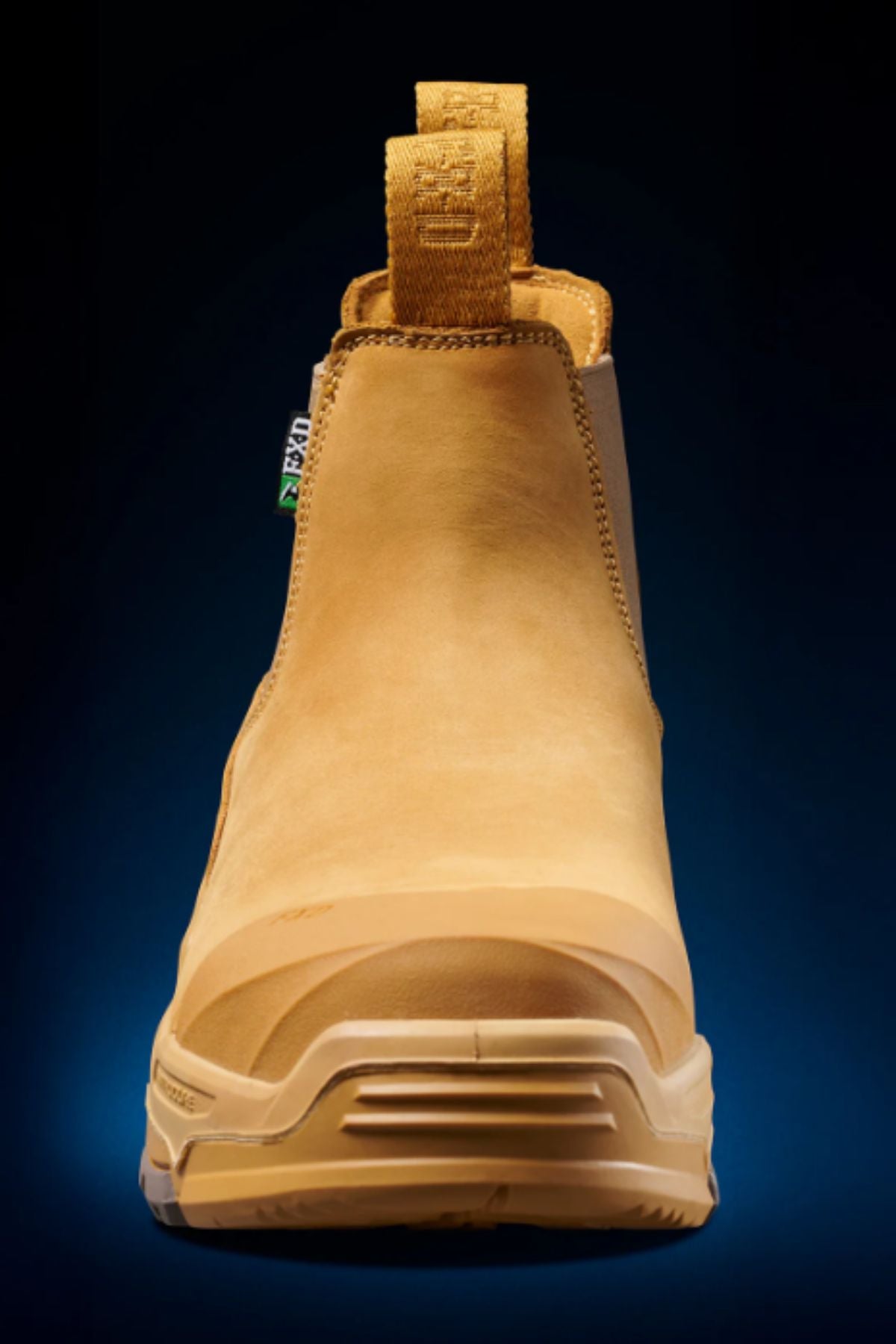 The FXD DURA X-3 Slip On Work Boots, featuring a tan color, pull tabs, and a durable puncture-resistant sole, are displayed from the front against a dark blue gradient background.
