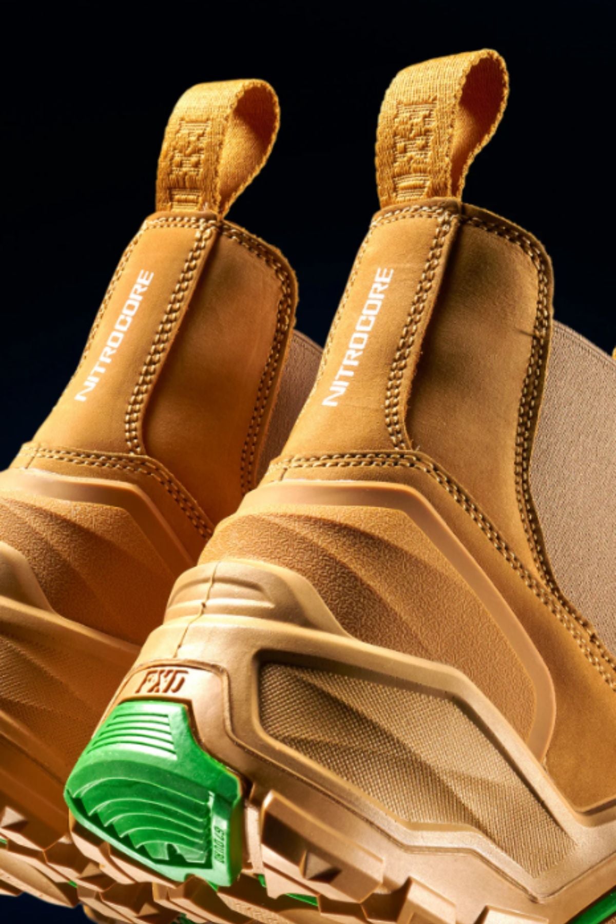 Close-up of FXD DURA X-3 Slip On Work Boots in tan, featuring rugged, puncture-resistant soles with green and white tread details and "NITRODORE" on the side pull-loops, set against a dark background.