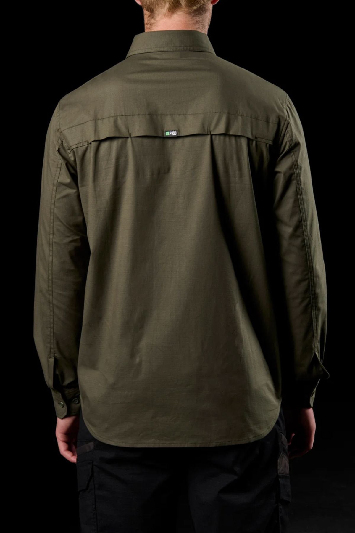 A person is seen from behind wearing an olive green FXD LSH-3 Vented Work Shirt with long sleeves and a collar, paired with black pants, against a black background.
