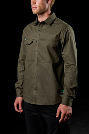 A man stands against a black background wearing the FXD LSH-3 Vented Work Shirt in olive green. The long-sleeve shirt features a collar, front pocket, subtle button detailing, and lightweight stretch fabric. Only his upper body is visible.