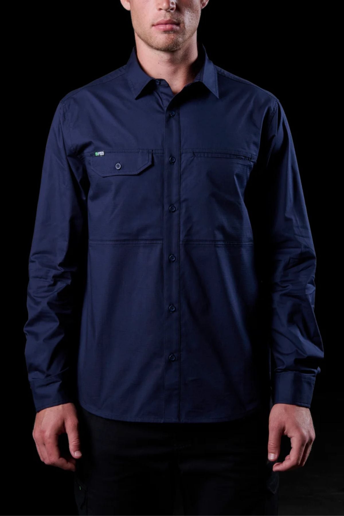 A man wearing the FXD LSH-3 Vented Work Shirt—long-sleeve, button-up, navy blue with chest pockets and lightweight stretch fabric—stands against a black background.