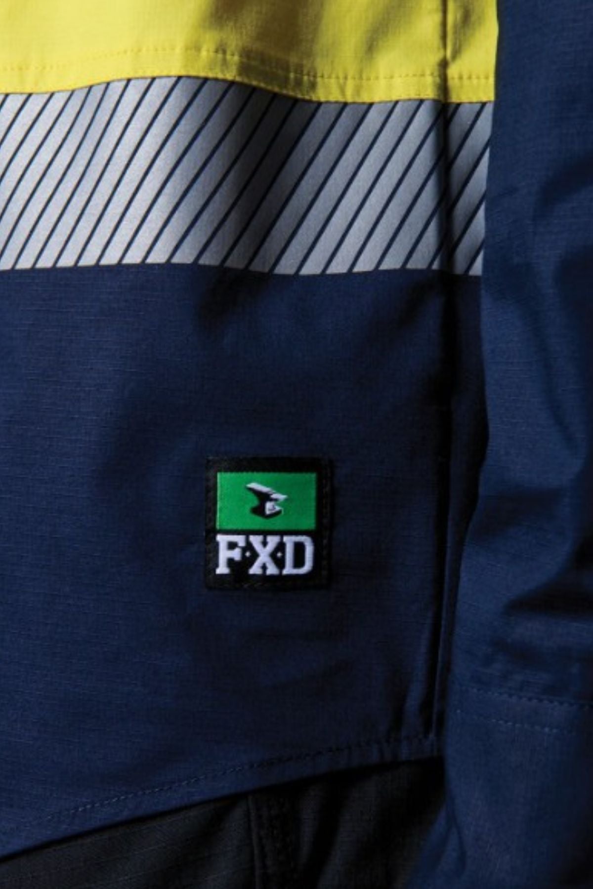 Close-up of the FXD LSH-4T Work Shirt, a blue and yellow shirt with segmented reflective tape, a diagonal white stripe, and a rectangular patch bearing "FXD" text on green with a white logo above.