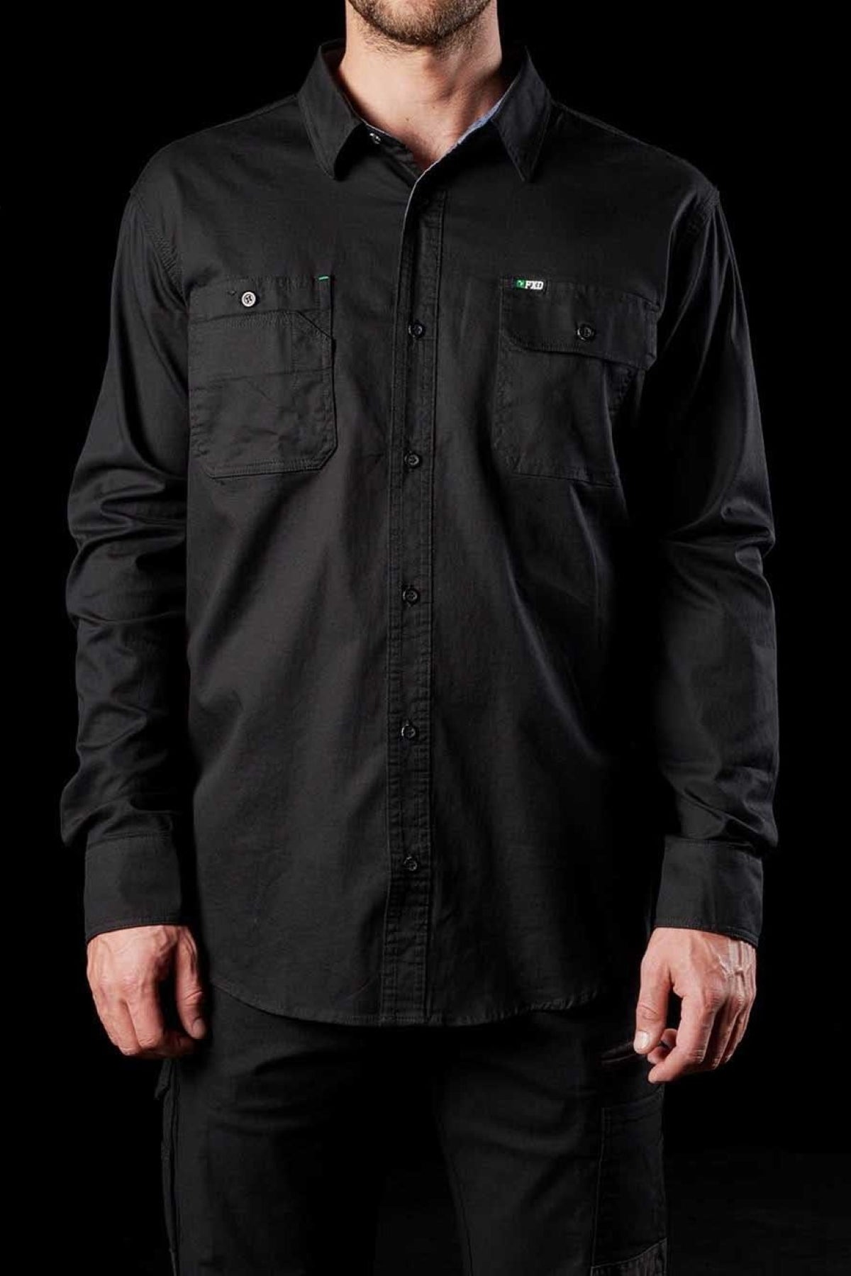 A person wearing the FXD LSH-1 Stretch Work Shirt, a long-sleeved black button-up with two chest pockets, stands against a black background. The durable shirt has a professional appearance, and the person's hands are at their sides.