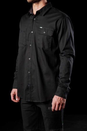 A man wearing an FXD LSH-1 Stretch Work Shirt with buttoned chest pockets stands against a black background. Only the torso and part of the arms are visible. The shirt has a casual, slightly loose fit, and the man's left hand is partially tucked into his pocket.