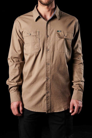 A person stands against a black background, wearing the FXD LSH-1 Stretch Work Shirt in tan. The long-sleeved shirt features durable triple needle seams and two chest pockets for added utility. They have paired the comfortable work shirt with dark pants. The individual's face is not visible in the image.