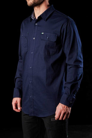 A man dressed in an FXD LSH-1 Stretch Work Shirt, navy blue and long-sleeved with two chest pockets. The shirt is adorned with a small embroidered logo on the left pocket and is designed for ultimate comfort and durability. Set against a black backdrop, only his torso and part of his arms are visible.