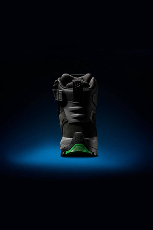 A single FXD WB-1 Work Boot in black is showcased against a dark background with a blue spotlight effect beneath it. The heel of the boot is prominently displayed, featuring a green sole and intricate design details on the upper, emphasizing its slip-resistant feature.