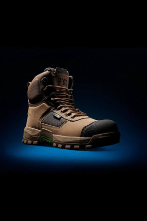 The FXD WB-1 Work Boot, a high-top, rugged tan work boot with distinctive black and green accents, is showcased against a dark background. This boot features robust construction with a thick, textured sole specifically designed for outdoor or heavy-duty use. Slip-resistant laces are tightly secured, underscoring its durability and safety.