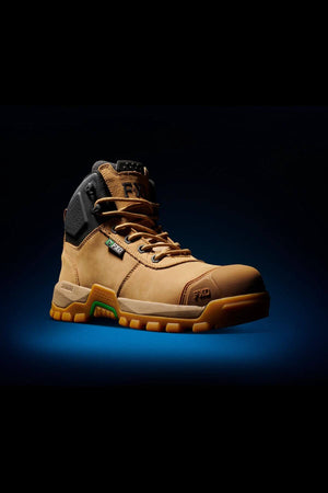 A single FXD WB-2 Work Boot, tan in color with a black padded collar, is displayed against a dark backdrop. The boot boasts a robust, slip-resistant sole with green accents and laces. It features safety toe reinforcement and includes logos and text highlighting its design for performance and safety.