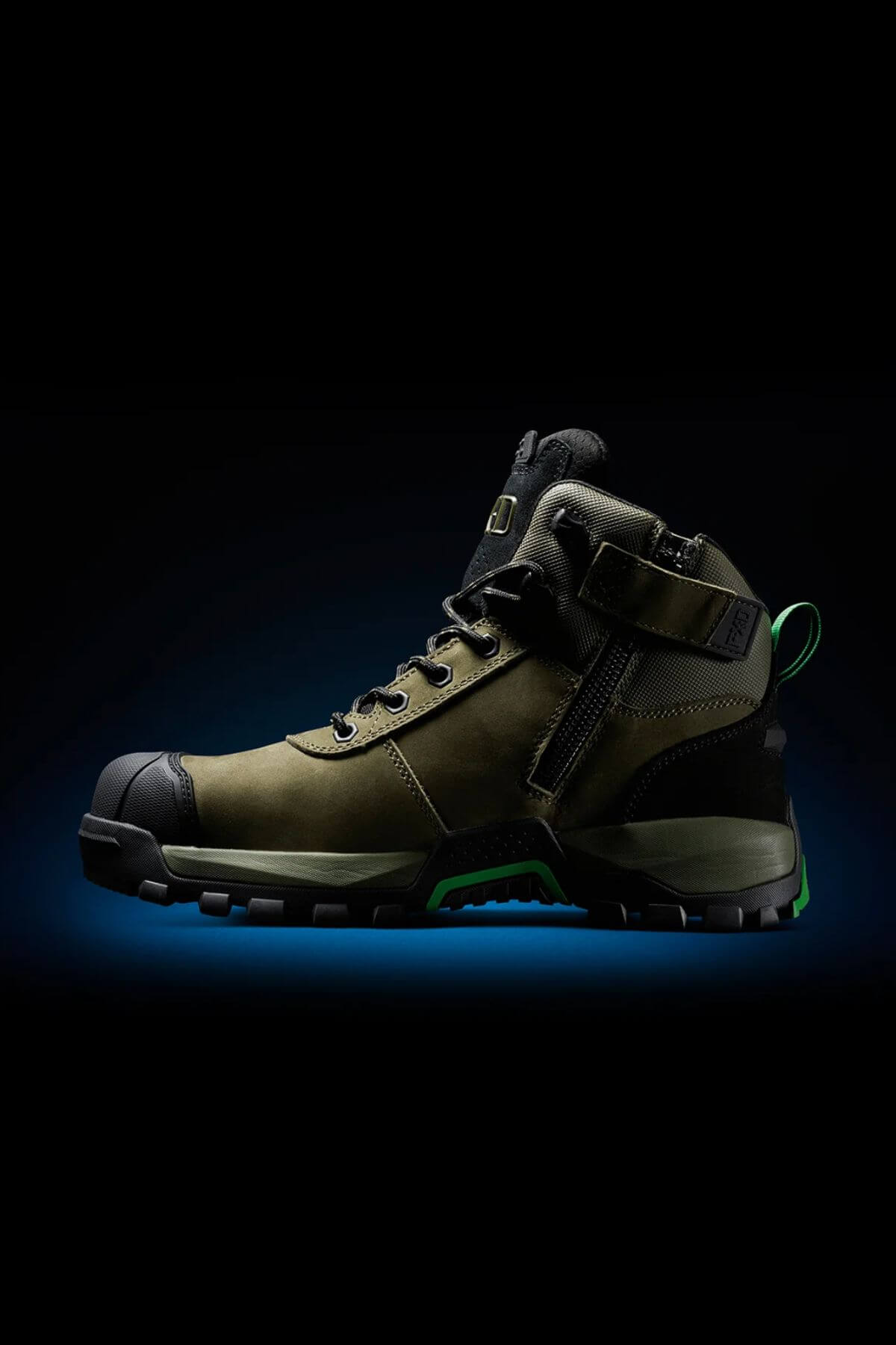The FXD WB-2 Work Boot is a rugged, olive green and black boot with a composite safety toe, thick sole, toe cap, side zipper, ankle strap, and NITROLITE midsole cushioning, displayed on a dark background with a blue glow.