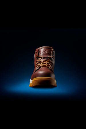 The FXD WB-3 Work Boot is a single brown premium leather boot with yellow laces and a tan sole, shown from the front on a dark blue gradient background with dramatic lighting.