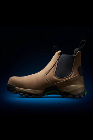 The FXD WB-4 Work Boot is a single brown slip-on boot featuring a black elastic side panel, a rugged black and green slip-resistant sole, and pull tabs at the front and back. It is displayed against a black background with a subtle blue glow around the base.