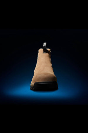 A single FXD WB-4 Work Boot in tan color, featuring slip-resistant soles, is showcased against a dark backdrop with a blue spotlight beneath it, emphasizing the shoe. The boot includes a pull tab at the back and exhibits stitching details around the ankle area.