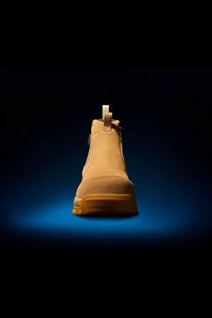 A single FXD WB-4 Work Boot in tan color, designed with a pull tab at the heel, is centered against a dark gradient blue and black background. The boot, equipped with a safety toe, faces forward to highlight its sturdy and durable design, detailed stitching, and thick sole.