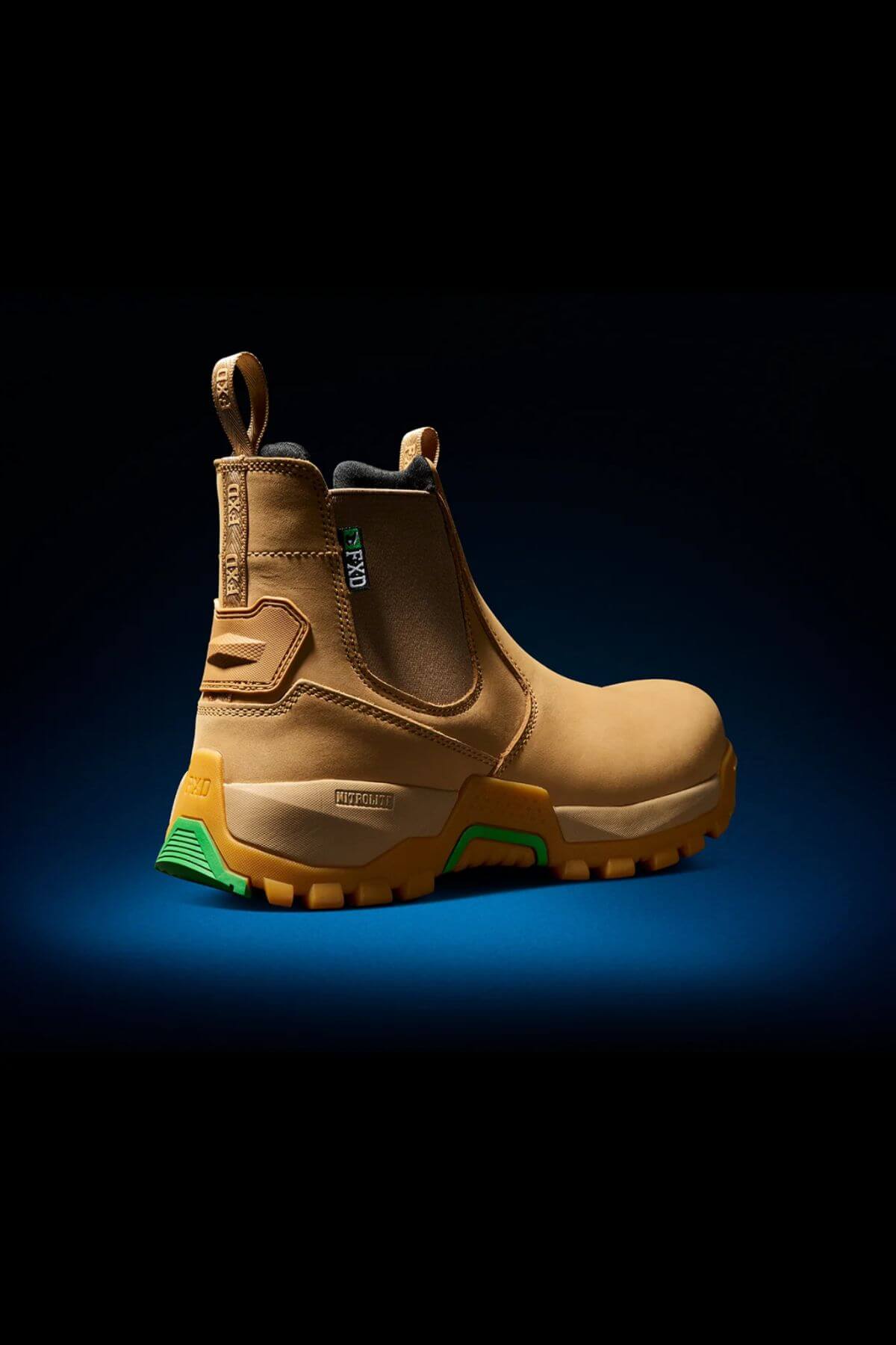 A single FXD WB-4 Work Boot in tan with green and yellow accents is centered against a dark blue background. The boot features a reinforced safety toe, pull-on tabs, and a rugged sole design. Additionally, the interior includes moisture-wicking properties to keep your feet dry all day.