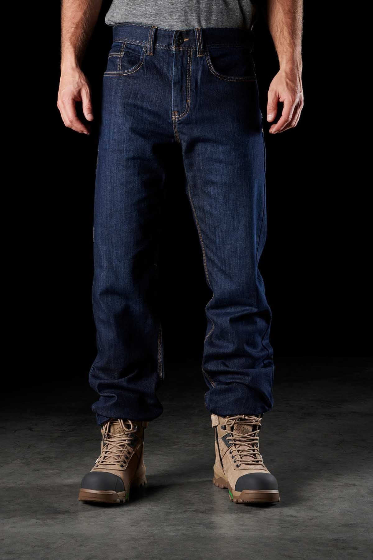 A person wearing FXD WD-2 blue denim jeans with DURA500™ reinforced pockets and tan work boots stands against a black background. The durable FXD WD-2 jeans are slightly loose-fitting, covering part of the boots. The person is also wearing a gray shirt that is partially tucked into the jeans.