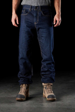 A person wearing FXD WD-2 blue denim jeans with DURA500™ reinforced pockets and tan work boots stands against a black background. The durable FXD WD-2 jeans are slightly loose-fitting, covering part of the boots. The person is also wearing a gray shirt that is partially tucked into the jeans.