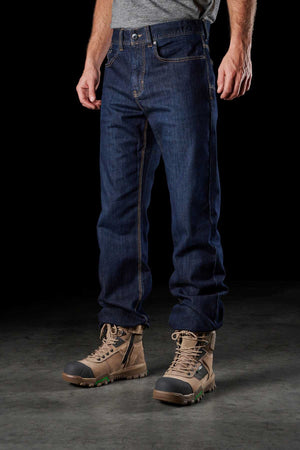 A person is standing against a dark background, wearing a gray shirt, FXD WD-2 blue jeans, and tan work boots. The classic fit jeans feature DURA500™ reinforced pockets for added strength, while the boots appear sturdy and designed for heavy-duty work. The photo focuses on the lower half of the individual.
