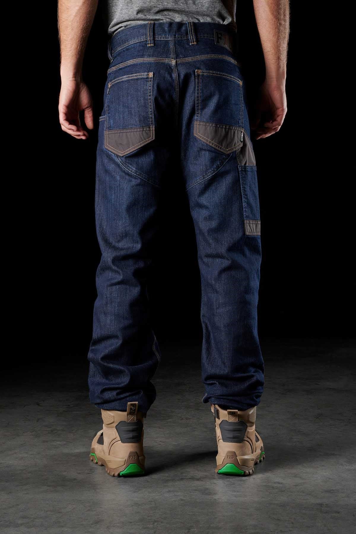 A person stands with their back facing the camera, wearing a pair of FXD WD-2 dark blue Superior Stretch S-GENE® denim jeans, which feature DURA500™ reinforced pockets and visible stitching for a casual fit. They are also sporting beige work boots with green soles. The dark background emphasizes the clothing.