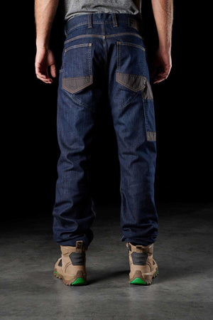 A person stands with their back facing the camera, wearing a pair of FXD WD-2 dark blue Superior Stretch S-GENE® denim jeans, which feature DURA500™ reinforced pockets and visible stitching for a casual fit. They are also sporting beige work boots with green soles. The dark background emphasizes the clothing.