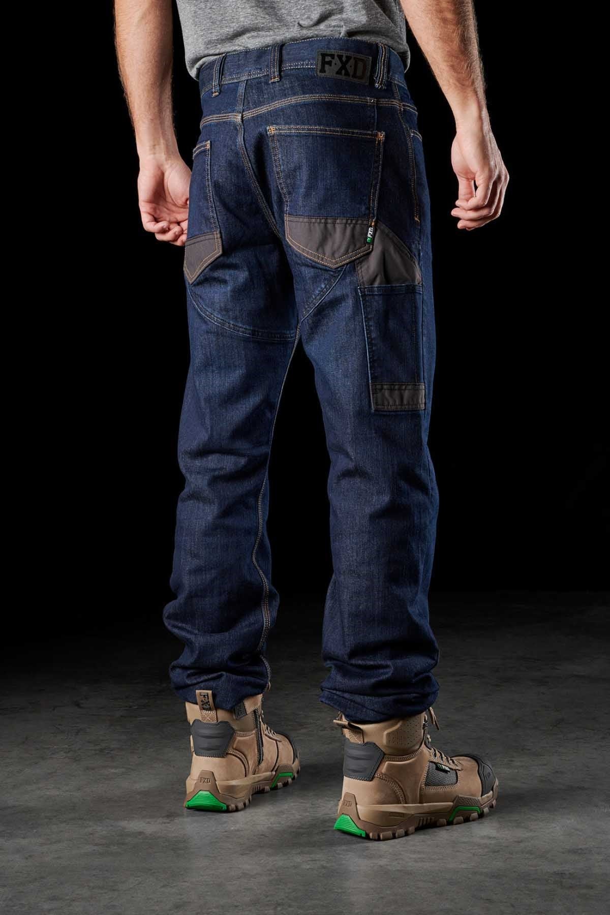 A person stands facing away from the camera, wearing a grey shirt, dark blue FXD WD-2 jeans featuring DURA500™ reinforced pockets and Superior Stretch S-GENE® denim, and tan work boots with green accents. The background is black, highlighting the durable clothing and footwear.