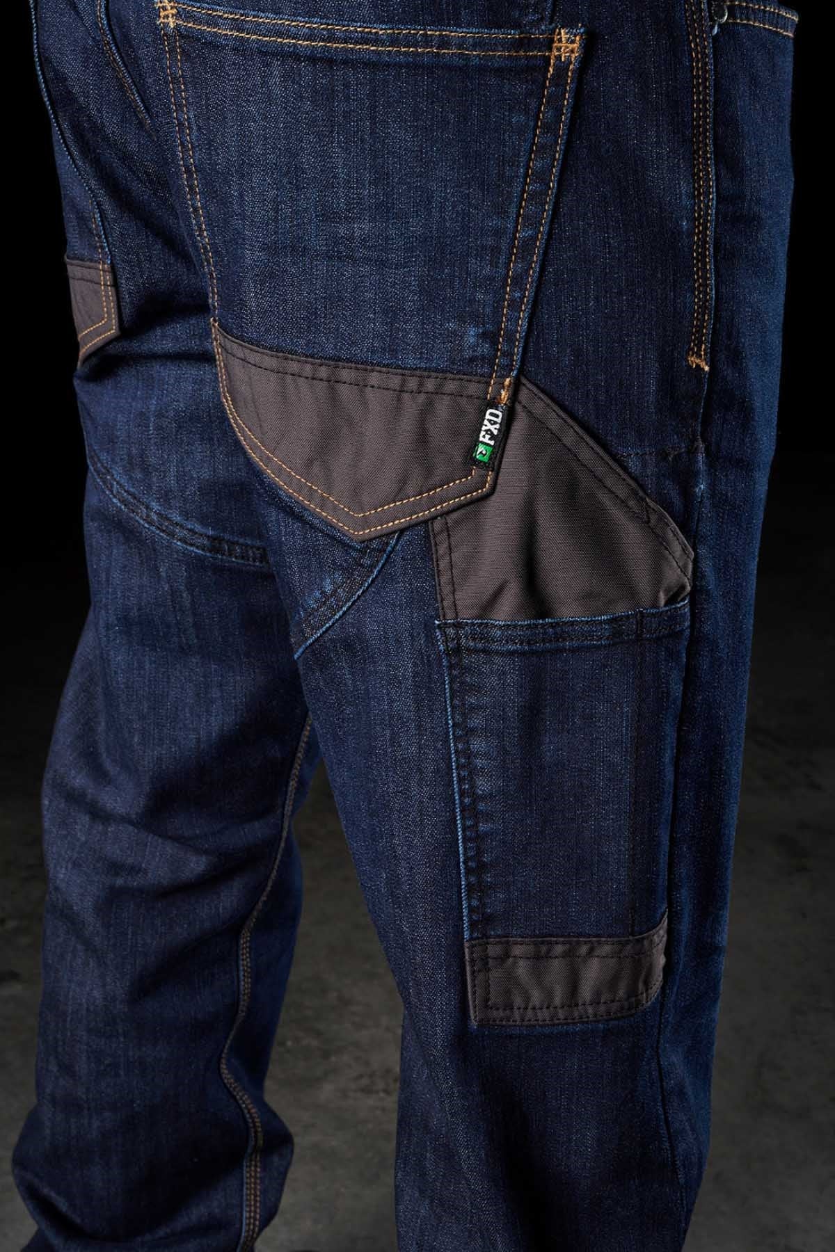 Close-up of a person's leg wearing the FXD WD-2 dark blue denim jeans, featuring multiple pockets, including a black fabric patch pocket. The DURA500™ reinforced pockets and prominent orange stitching enhance durability, while a small green label near one of the pockets catches the eye. The dark background emphasizes the jeans.