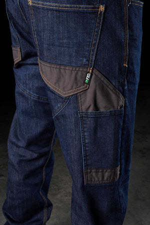 Close-up of a person's leg wearing the FXD WD-2 dark blue denim jeans, featuring multiple pockets, including a black fabric patch pocket. The DURA500™ reinforced pockets and prominent orange stitching enhance durability, while a small green label near one of the pockets catches the eye. The dark background emphasizes the jeans.