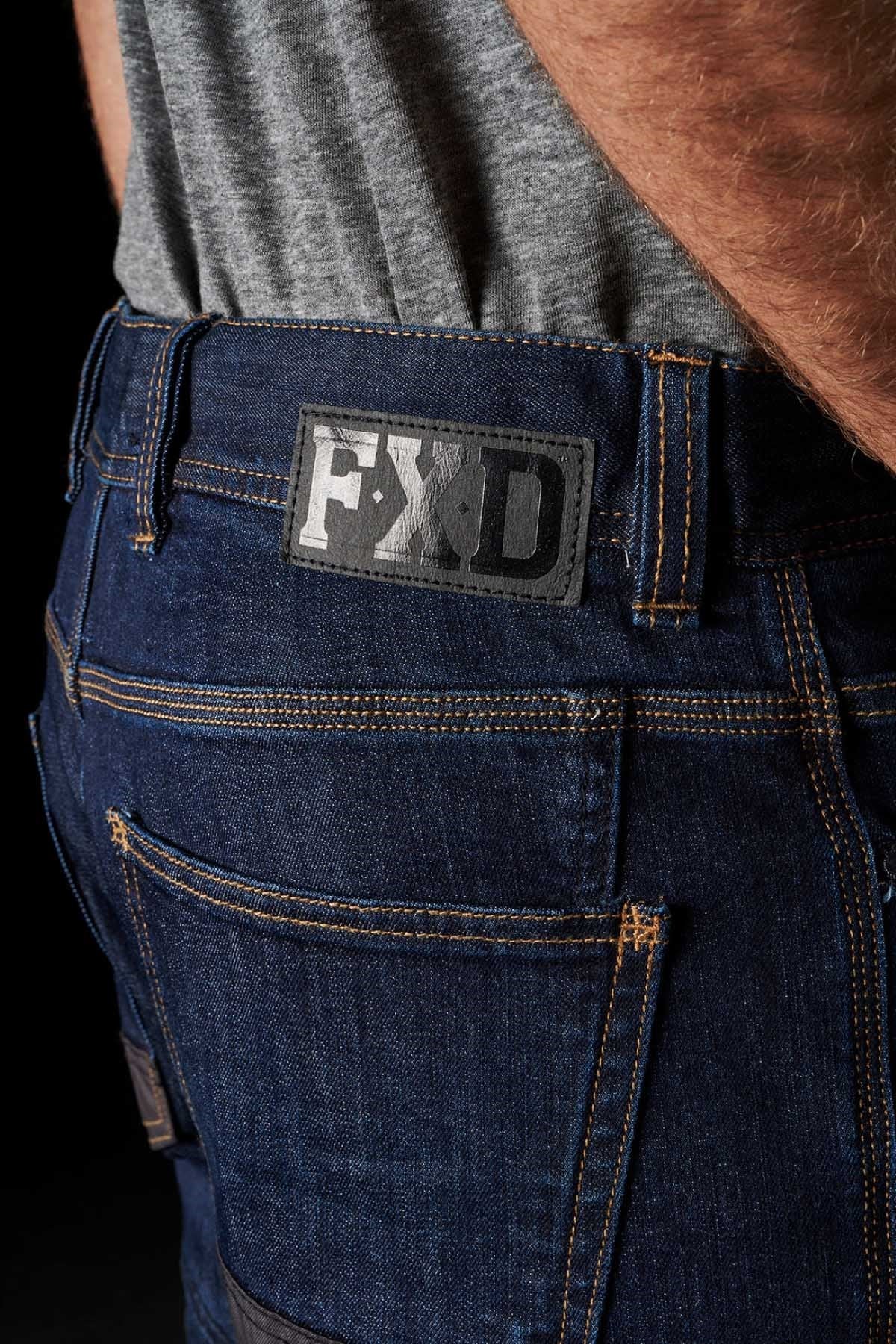 Close-up of a person wearing dark blue FXD WD-2 Superior Stretch S-GENE® denim jeans with visible stitching details. The jeans feature a black leather patch on the waistband with the "FXD" brand logo and DURA500™ reinforced pockets. The person is also wearing a grey shirt tucked into the jeans.