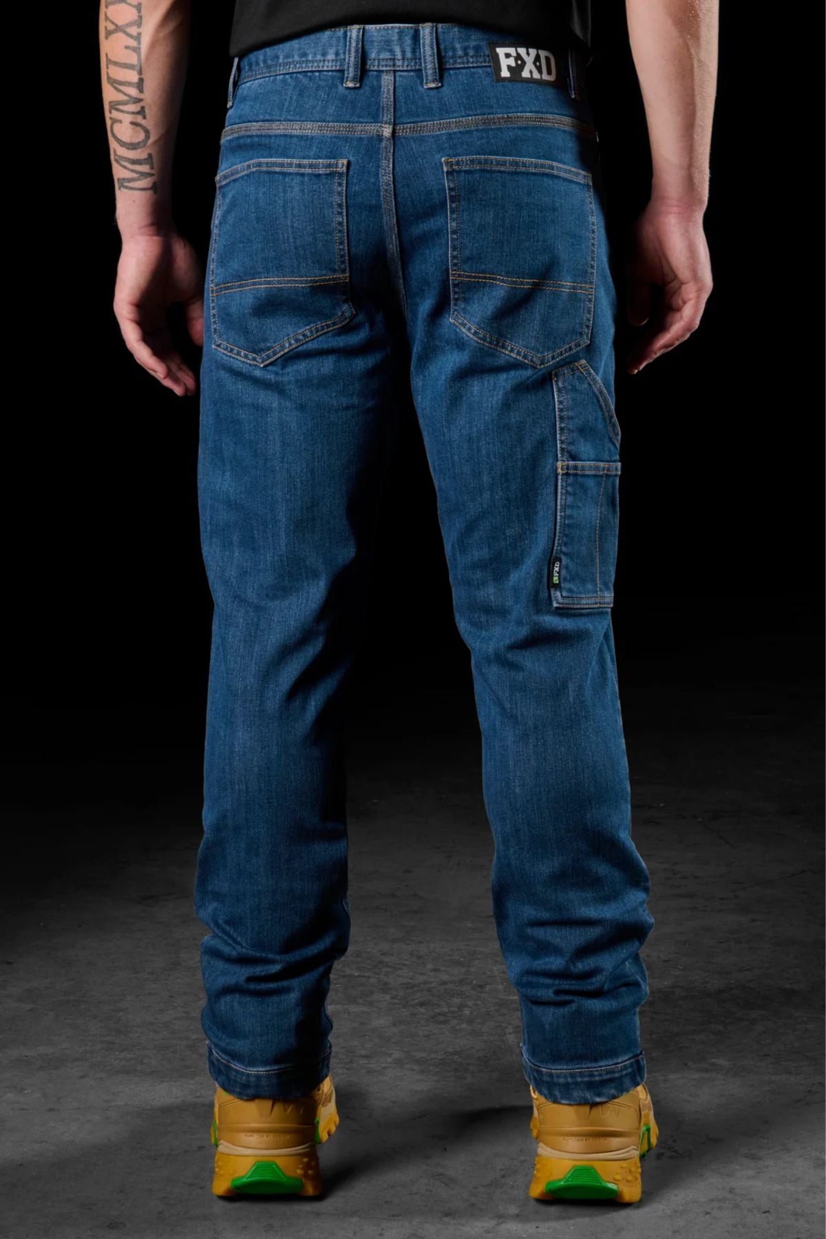 Someone wearing FXD WD-4 jeans made from COOLMAX EcoMade fabric and yellow shoes stands facing away, arms relaxed, displaying the back pockets and a tattoo on their right arm against a dark backdrop.