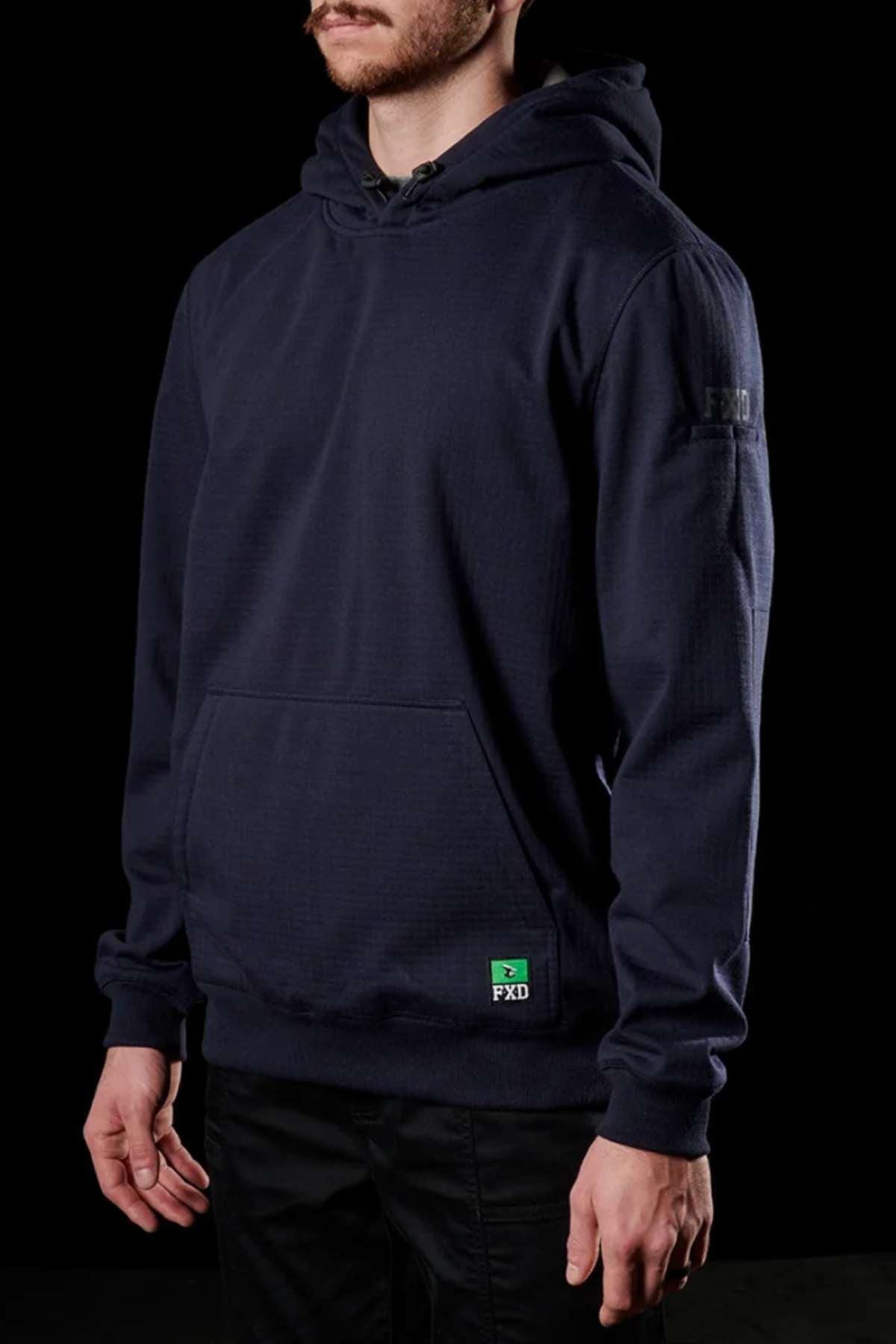 A person wearing a dark navy FXD WF-1 Hoodie stands against a black background. The windproof fleece hoodie features a kangaroo pocket in the front and a small green FXD logo patch near the bottom. The person's face is not visible.