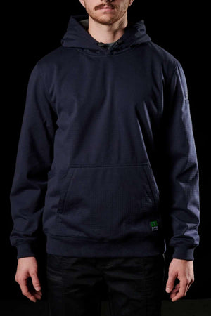 A man is standing against a black background wearing a dark navy FXD WF-1 Hoodie with a front pocket and black pants. The hoodie, made from windproof fleece, features a small green embroidered patch near the pocket. The man's face is partially visible, highlighting the clothing.