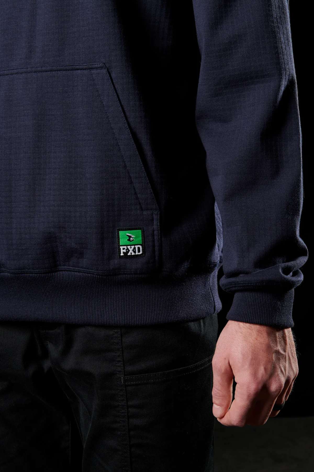 A person stands with their right arm relaxed at their side, wearing a dark navy FXD WF-1 Hoodie and black pants. The corner of a front pocket and a small green and black FXD logo are visible on the water-resistant hoodie. Only part of the person's torso and hand are shown.