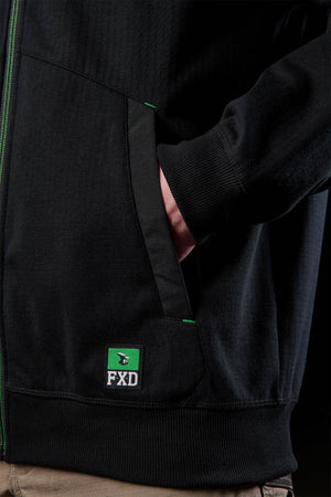 Wearing a black FXD WF-3 Jacket with green accents and the FXD logo near the hem, a person stands with their hand in the pocket. The water-resistant jacket is styled with tan workwear pants for a practical look.