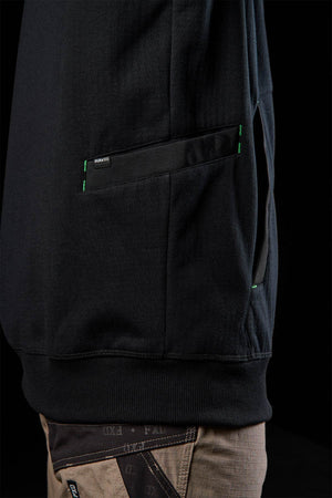 A close-up of a person wearing the FXD WF-3 Jacket, a black workwear-inspired sweatshirt with green pocket stitching, paired with tan pants featuring black and brown patches, set against a black background.