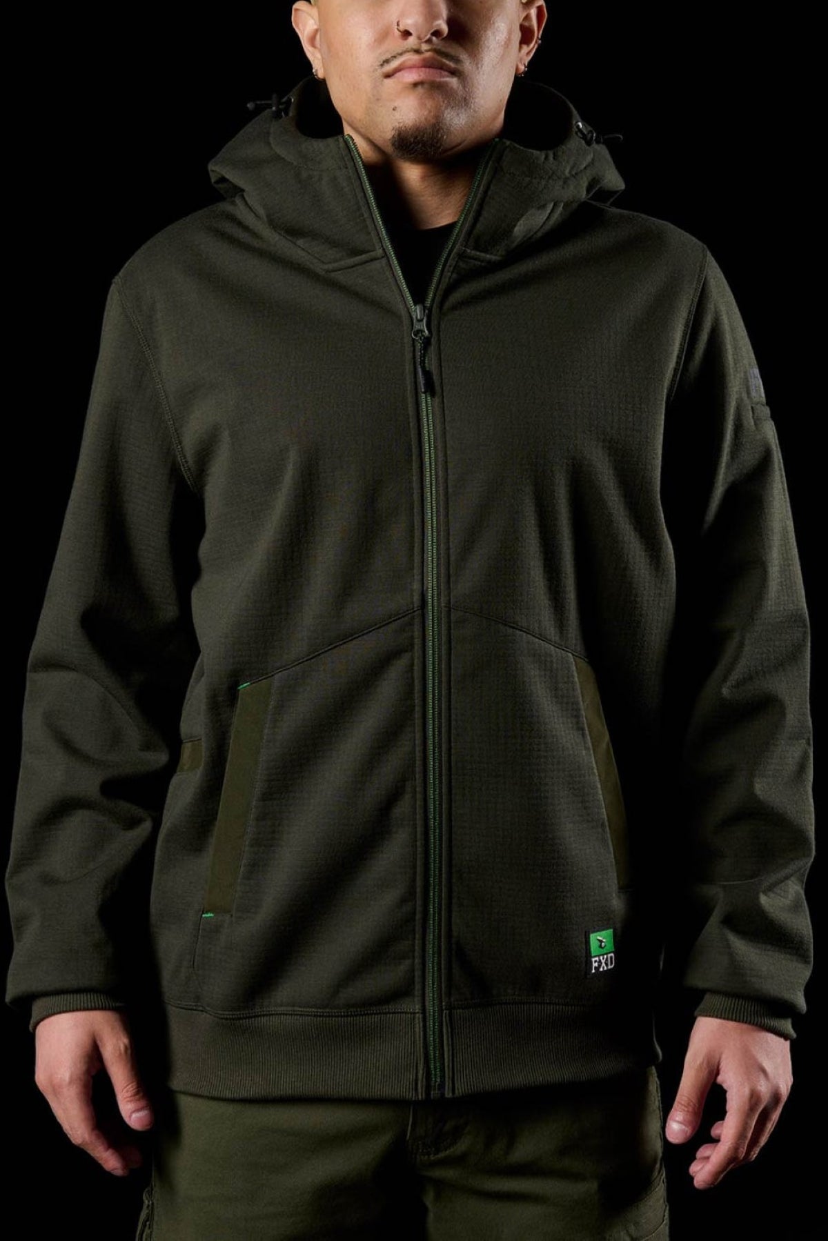 A person in a dark green FXD WF-3 Jacket with green pockets stands against a black background. Their face is partly visible and their hands relaxed, highlighting the stylish, water-resistant FXD WF-3 Jacket.