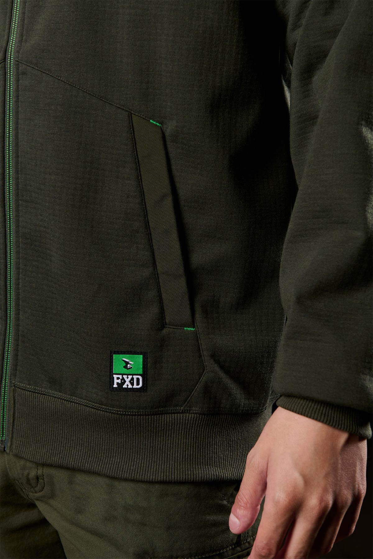 A person wearing a dark green FXD WF-3 Jacket and matching pants, with their right hand by their side. The jacket features a front pocket, an FXD logo patch near the pocket, and water-resistant design.