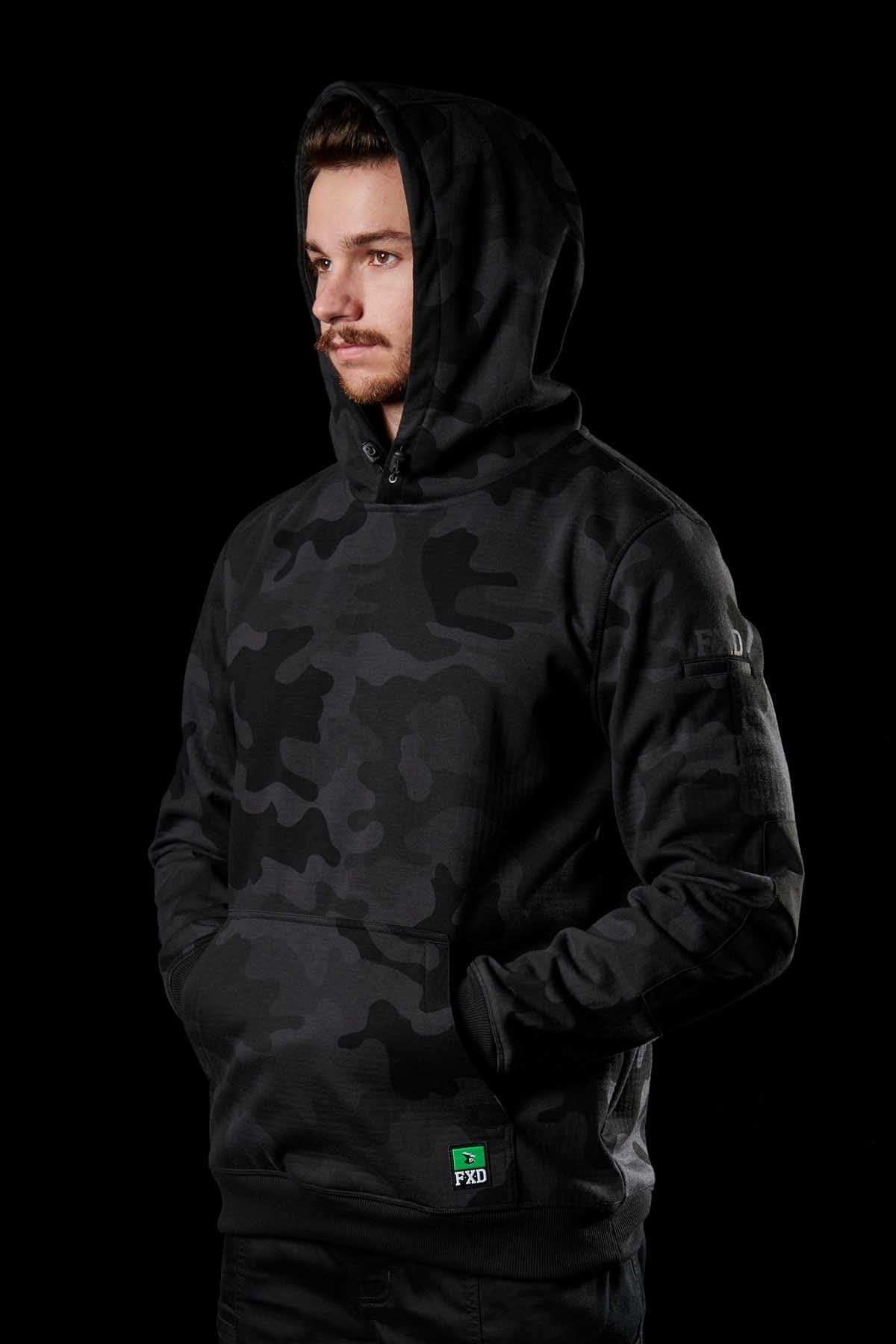 A person stands facing slightly to the right, wearing a dark camouflage patterned FXD WF-1 Hoodie with the hood up against a black background. The person's hands are in the front pockets of the windproof DURA500™ canvas hoodie, and a small green label with "FXD" is visible near the hem.