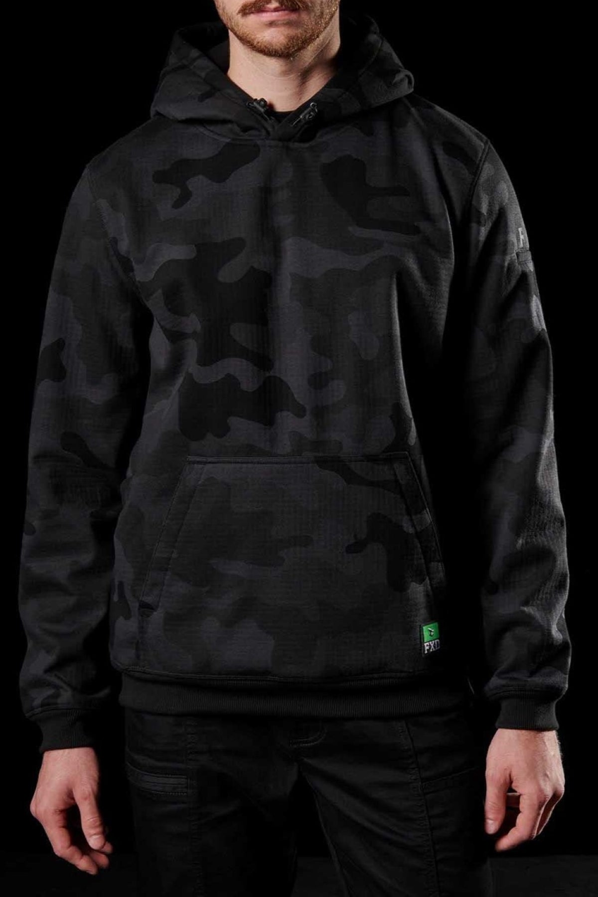 A person is wearing an FXD WF-1 Hoodie in black and gray camouflage, featuring a front pocket with a small green tag, paired with black pants. The dark background accentuates the individual's attire, making it the focal point of the image.
