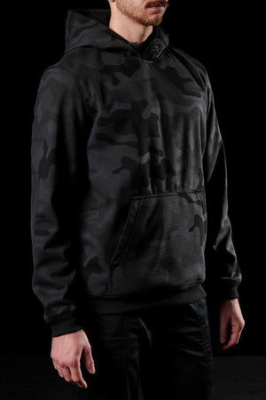 A person wearing an FXD WF-1 Hoodie stands against a black background. Only the lower part of their face is visible, and their hands are relaxed at their sides. The black windproof hoodie features a front pocket and a drawstring for adjusting the hood.