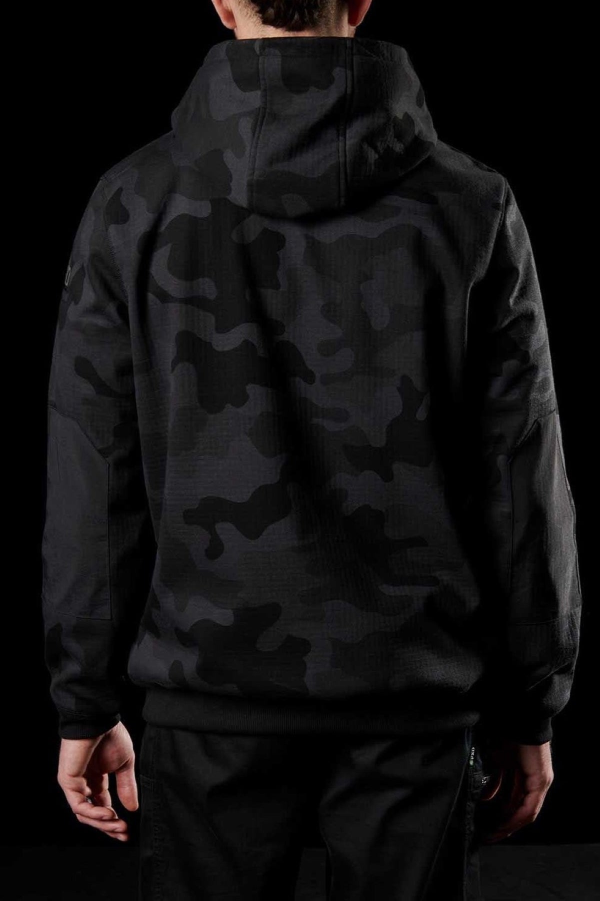 A person wearing an FXD WF-1 Hoodie made of black DURA500™ canvas with a dark camouflage pattern is seen from the back. They are also wearing black pants. The completely black background makes their clothing blend in slightly.