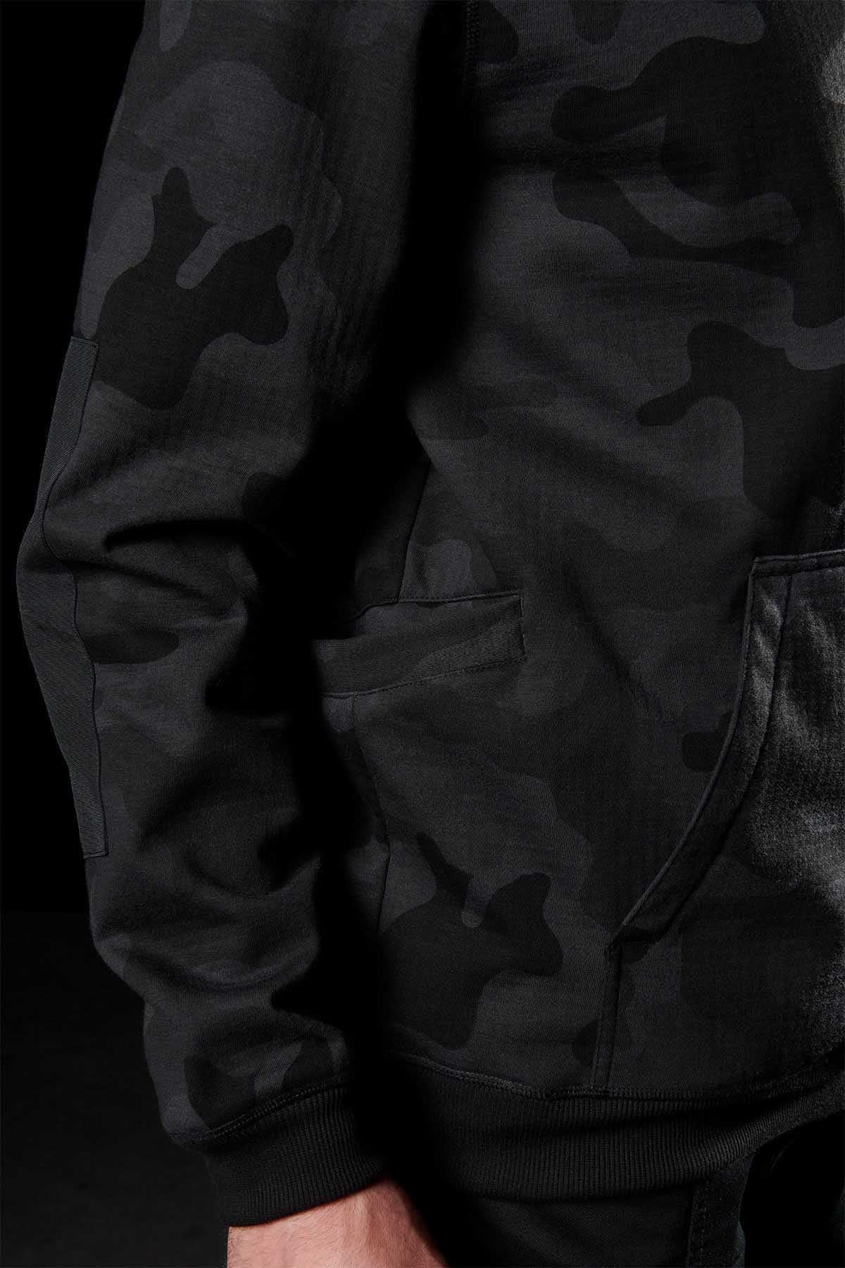 Close-up of a person wearing a black and dark gray camouflaged FXD WF-1 Hoodie. The hoodie features a front pocket, and the person's hand is partially visible at the bottom of the image. The background is dark.
