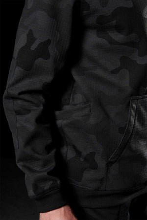 Close-up of a person wearing a black and dark gray camouflaged FXD WF-1 Hoodie. The hoodie features a front pocket, and the person's hand is partially visible at the bottom of the image. The background is dark.