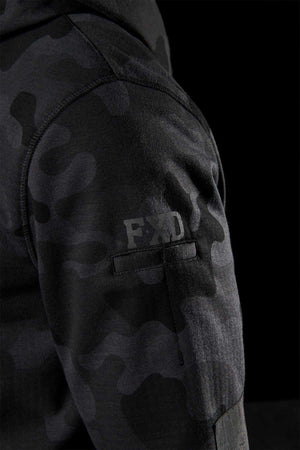 Close-up of a person's arm wearing an FXD WF-1 Hoodie in a black and gray camouflage pattern, with "FXD" printed on the sleeve, crafted from DURA500™ canvas. The background is completely black.