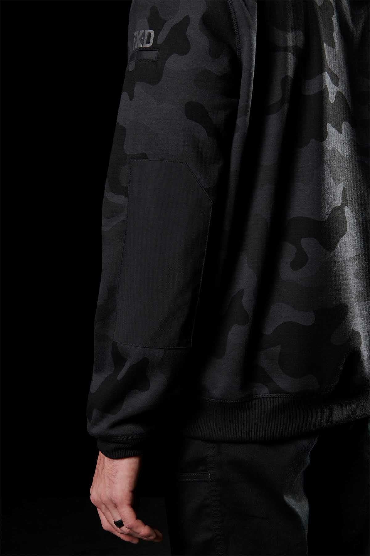 A person wearing an FXD WF-1 Hoodie in a dark camouflage pattern with a wrist patch and ribbed detailing on the sleeves stands against a black background. The left side of the torso and part of the right arm and hand are visible, showcasing the quality of DURA500™ canvas material.
