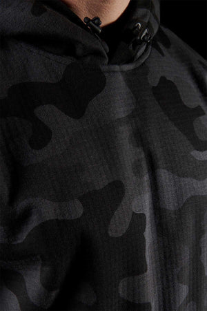 Close-up of a person wearing the FXD WF-1 Hoodie, a dark camouflage, windproof hoodie. The fabric showcases various shades of black and gray in irregular patterns. The hood is cinched at the neck with black drawstrings set against a matching black background.