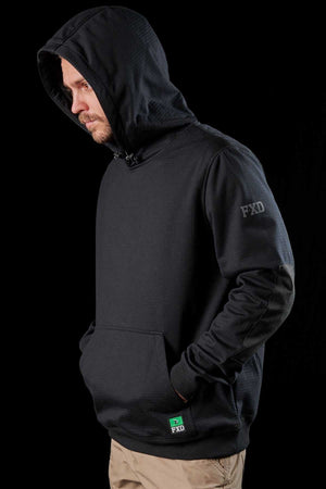 A man stands against a black background wearing an FXD WF-1 Hoodie with the hood up, showcasing the "FXD" logo on the sleeve and pocket, paired with beige pants. The windproof and water-resistant fleece hoodie has his hands tucked into its front pocket, and he sports stubble on his face while appearing to look downwards.