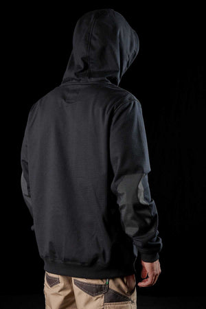 A person stands against a black background, viewed from behind. They are wearing the FXD WF-1 Hoodie with the hood up and beige pants. The individual's head is tilted down slightly, showcasing the DURA500™ canvas elbow patches on their sleeves, and their left hand is visible by their side.