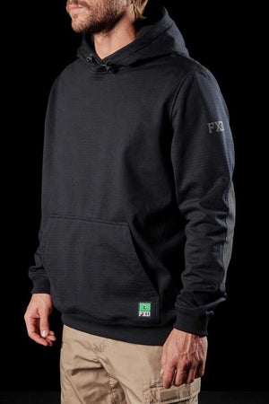 A bearded individual is dressed in a black FXD WF-1 Hoodie with the FXD logo displayed on the chest and sleeve, featuring durable DURA500™ canvas elbow patches. They are also sporting beige pants. The black background emphasizes their outfit.