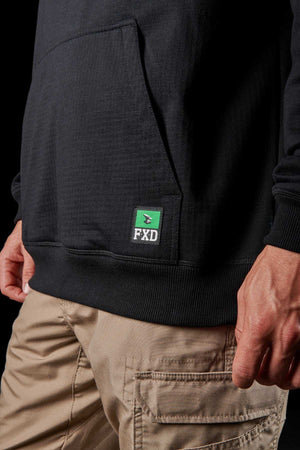 A person wearing the black FXD WF-1 Hoodie with DURA500™ canvas elbow patches and beige cargo pants stands with their left hand resting on the side of their pants. The hoodie features a green logo with white text reading "FXD" near the front pocket. The background is black.