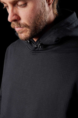 A close-up image portrays a man with short hair and a beard, gazing downward. He is dressed in an FXD WF-1 Hoodie with the hood down, highlighted by DURA500™ canvas elbow patches. The dimly lit background accentuates the man's profile and the hoodie’s fabric texture.